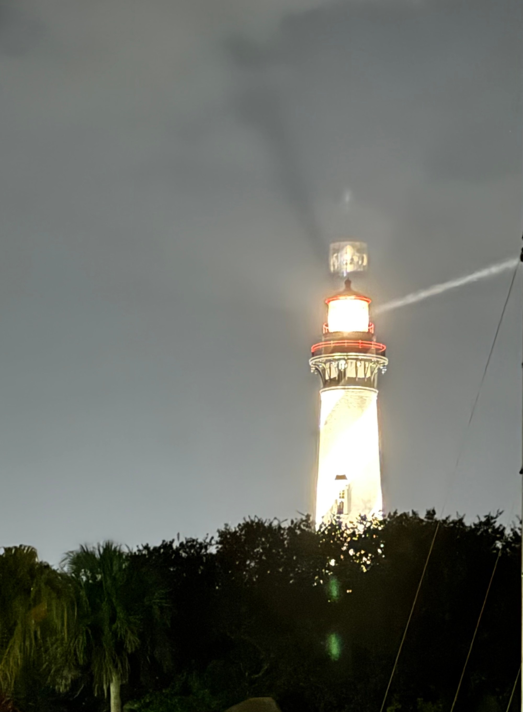 PP.08.25.11 Lighthouse illuminated at night, surrounded by trees, under a cloudy sky.